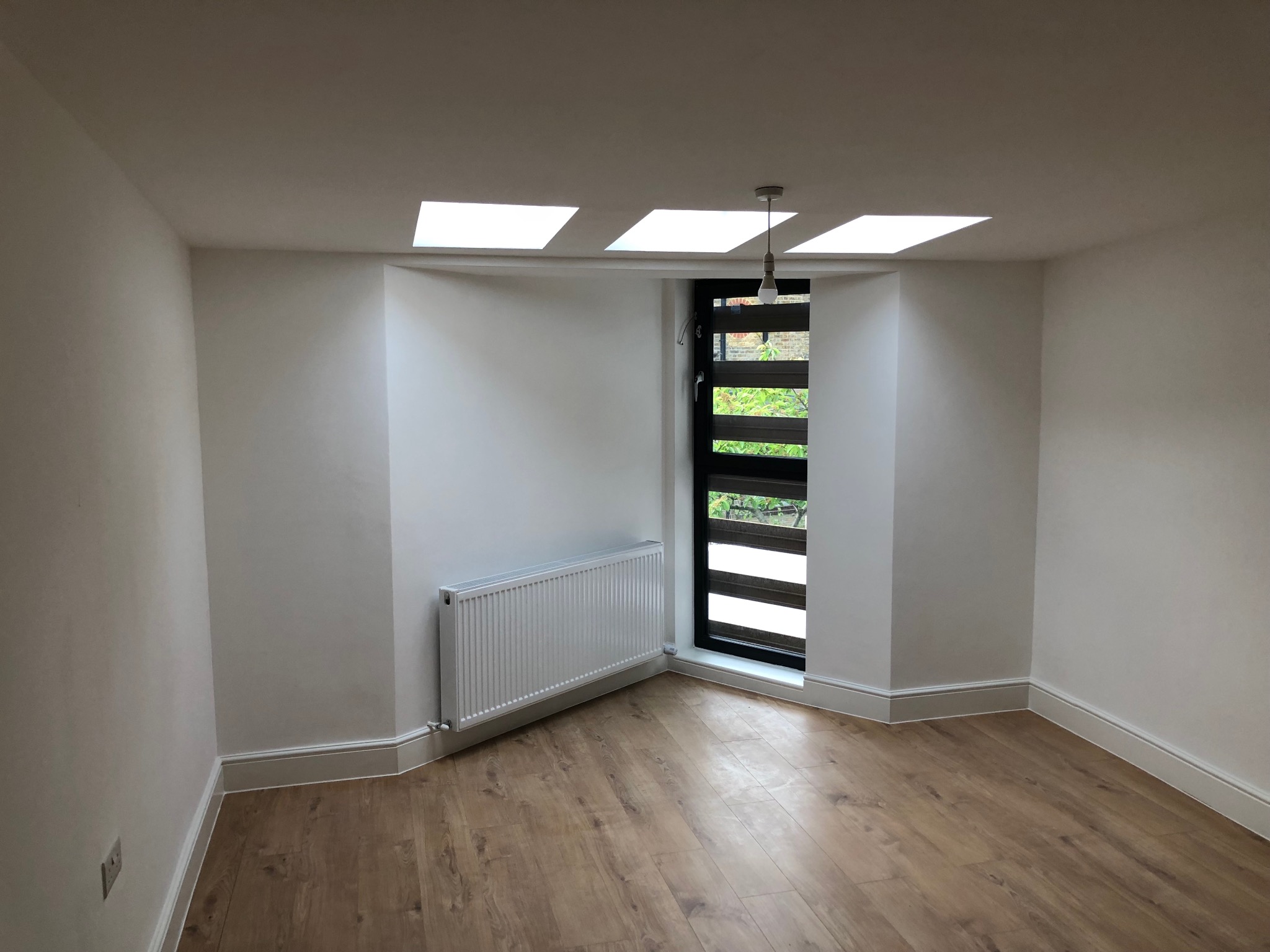 Wnętrze pokoju po remoncie: świeżo położone panele podłogowe, białe ściany z grzejnikiem i oknem z żaluzjami, widoczne trzy okna dachowe.