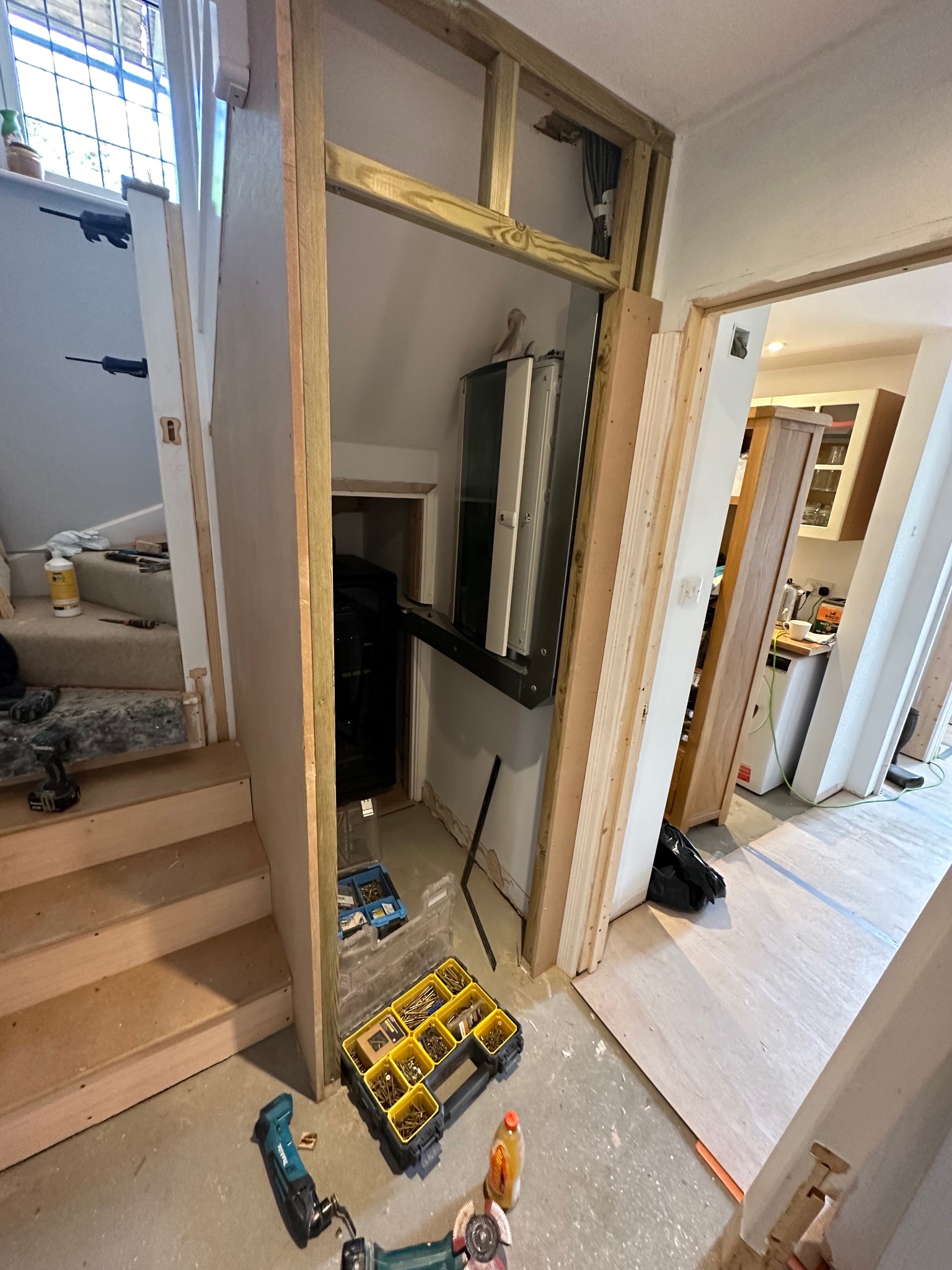 Adaptacja przestrzeni pod schodami: konstrukcja szkieletowa z drewna, narzędzia i materiały budowlane na podłodze, widok na korytarz i kuchnię w tle.