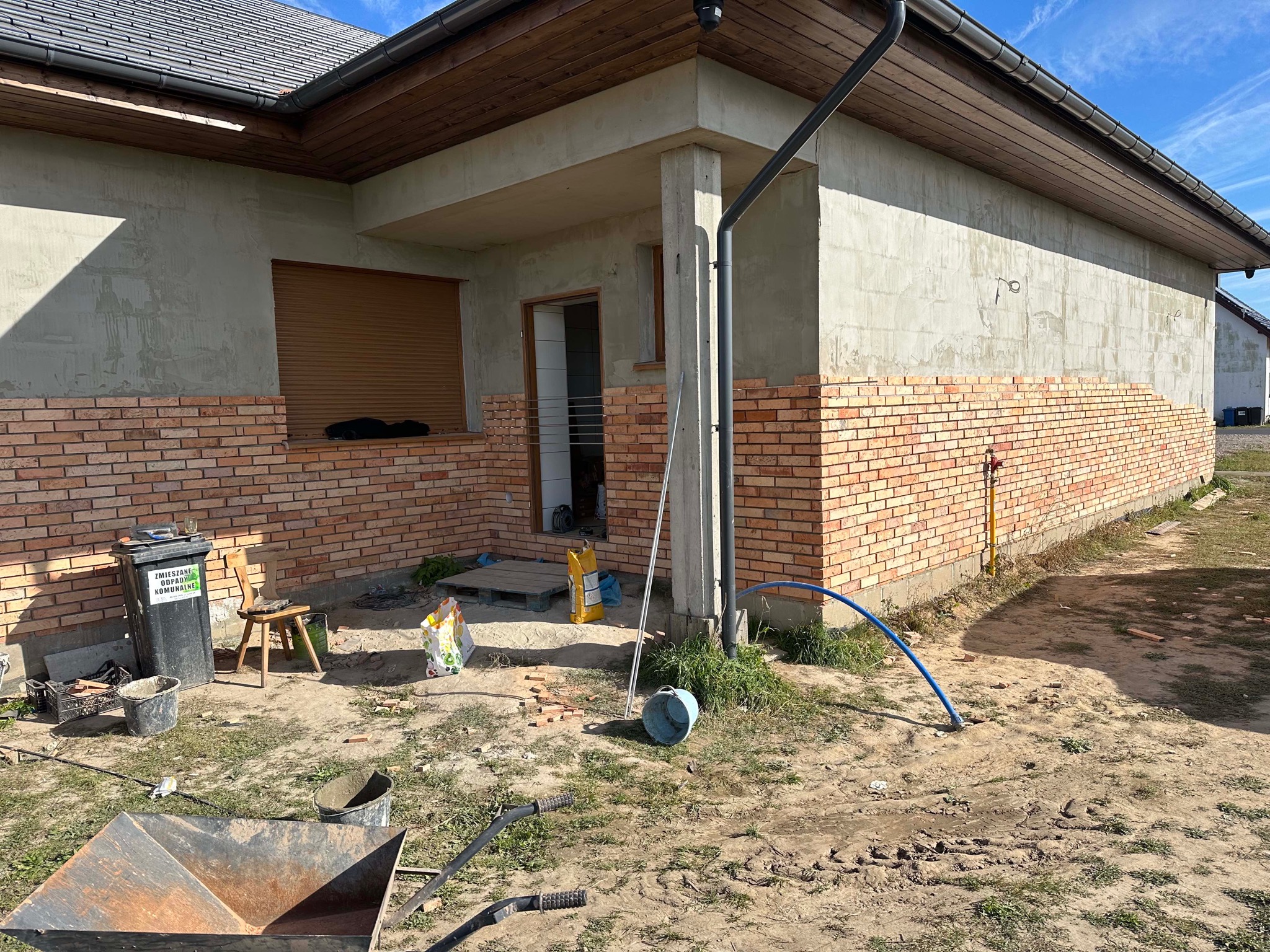 Montaż cegieł klinkierowych na elewacji domu jednorodzinnego, widoczne narzędzia i materiały budowlane, częściowo otynkowana ściana, przygotowana pod dalsze prace.