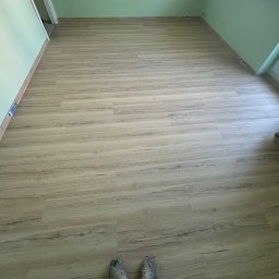 Well Done Krystian Chojnicki - Jasne panele podłogowe ułożone w pokoju z zielonymi ścianami, widoczne fragmenty butów w dolnej części kadru.