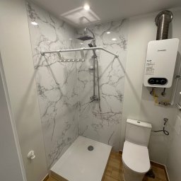 Well Done Krystian Chojnicki - Nowoczesna kabina prysznicowa z panelami imitującymi marmur, biały brodzik i armatura, wiszący piec gazowy Junkers oraz toaleta w małej łazience z drewnopodobną podłogą.