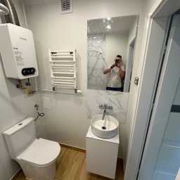 Well Done Krystian Chojnicki - Nowoczesna łazienka z białymi ścianami, wiszącą umywalką na szafce, podgrzewaczem wody Junkers, grzejnikiem drabinkowym i lustrem odbijającym osobę robiącą zdjęcie.