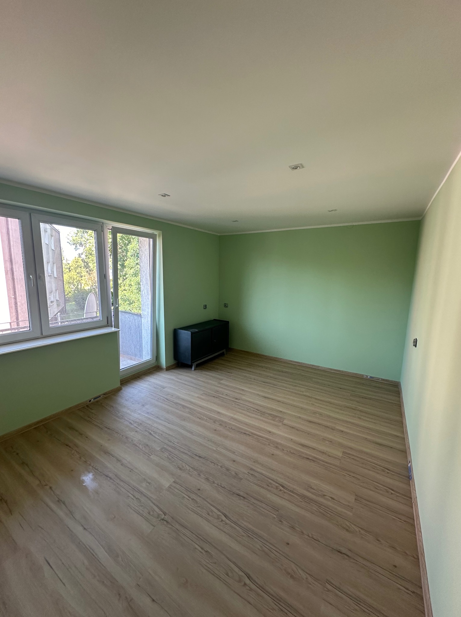 Pomalowane na jasnozielono puste pomieszczenie z panelami podłogowymi, oknem balkonowym i czarną szafką pod ścianą