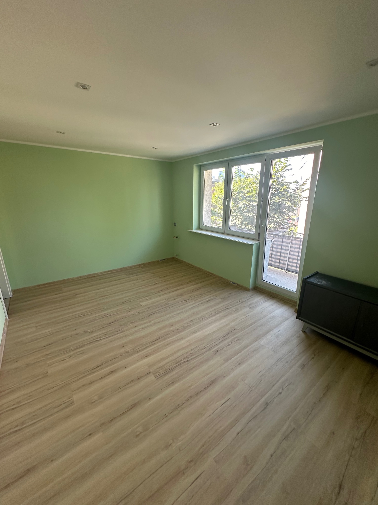 Pomalowane na pistacjowo wnętrze z panelami podłogowymi, białymi listwami przypodłogowymi i oknem balkonowym z widokiem na zieleń.