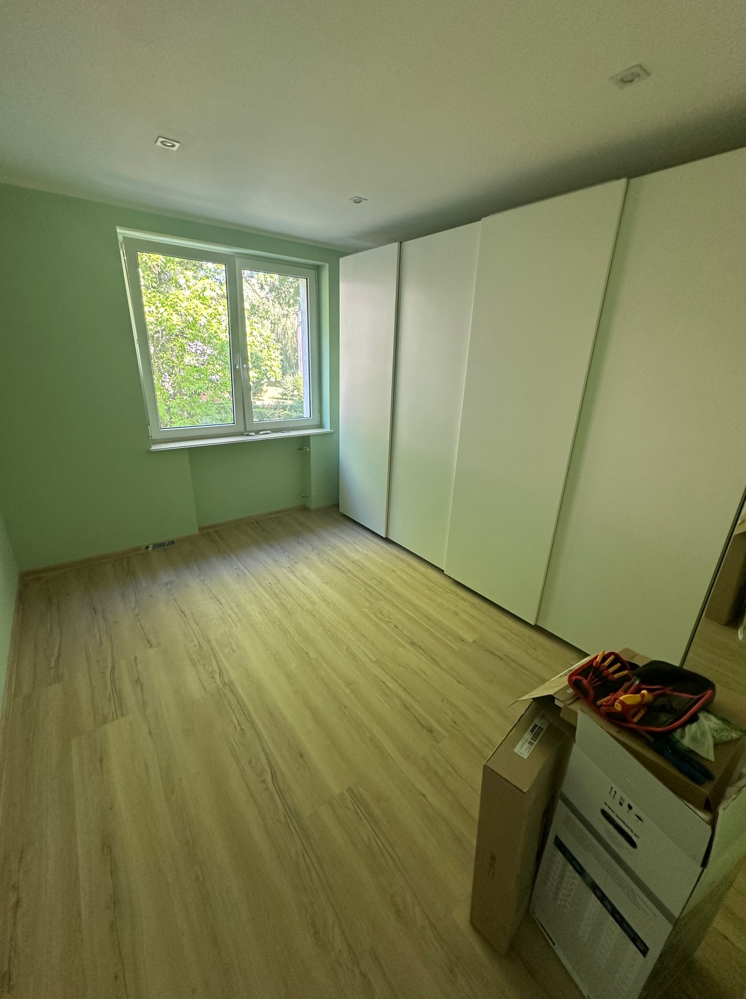 Pokój z zielonymi ścianami, jasną podłogą, białymi szafami wnękowymi i oknem z widokiem na drzewa; na podłodze kartony i narzędzia.