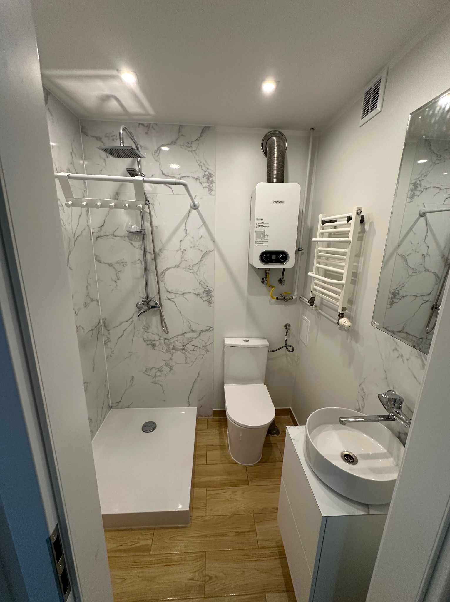 Nowoczesna, wąska łazienka z kabiną prysznicową, umywalką nablatową, toaletą i podgrzewaczem wody, wykończona płytkami imitującymi marmur i drewnopodobnymi.