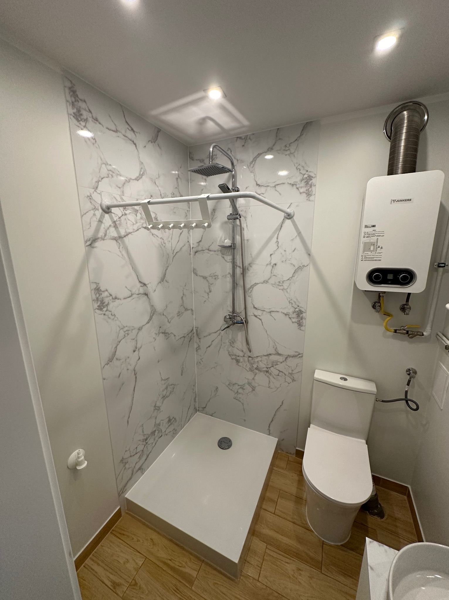 Nowoczesna kabina prysznicowa z panelami imitującymi marmur, biały brodzik i armatura, wiszący piec gazowy Junkers oraz toaleta w małej łazience z drewnopodobną podłogą.