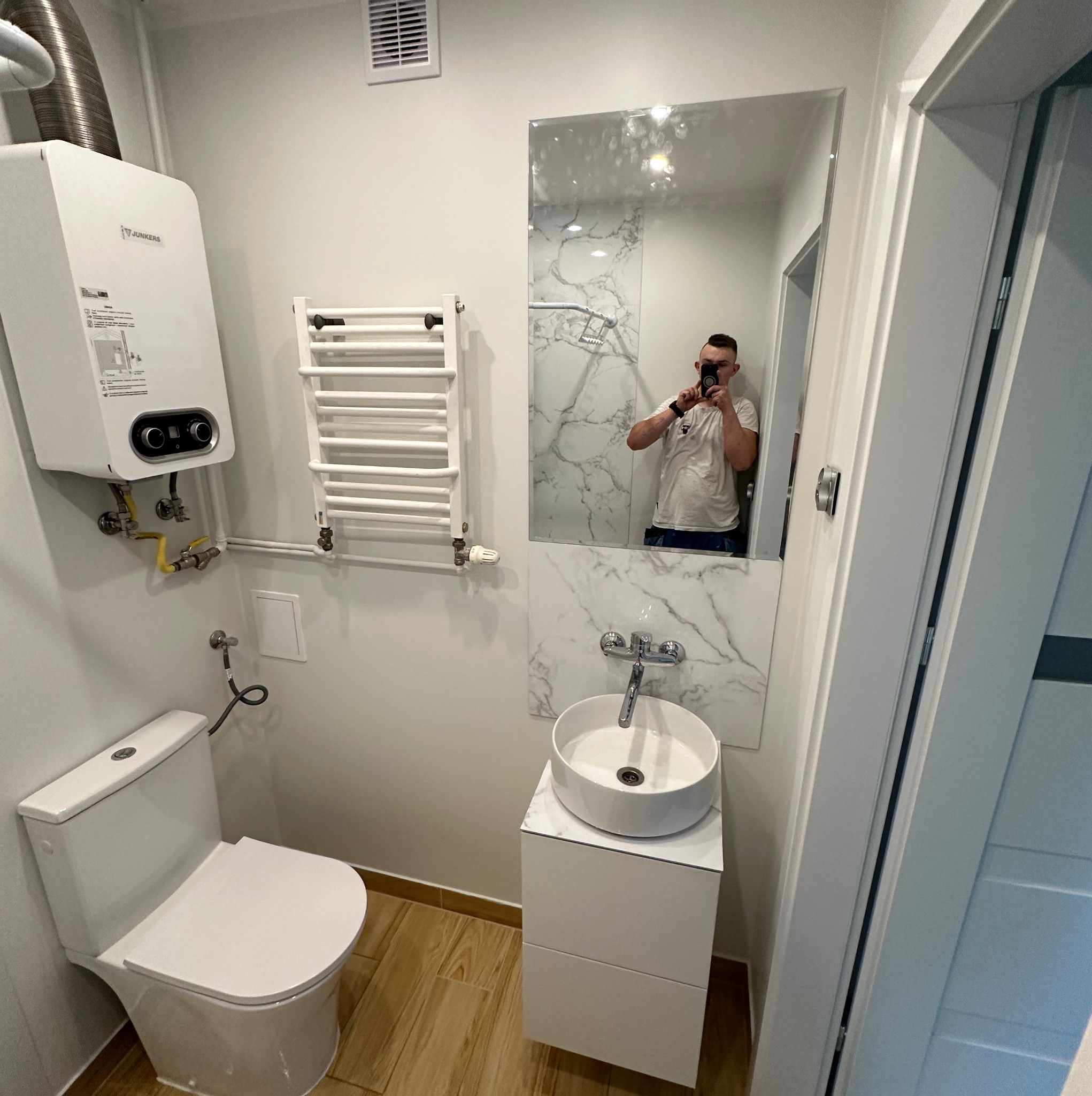 Nowoczesna łazienka z białymi ścianami, wiszącą umywalką na szafce, podgrzewaczem wody Junkers, grzejnikiem drabinkowym i lustrem odbijającym osobę robiącą zdjęcie.