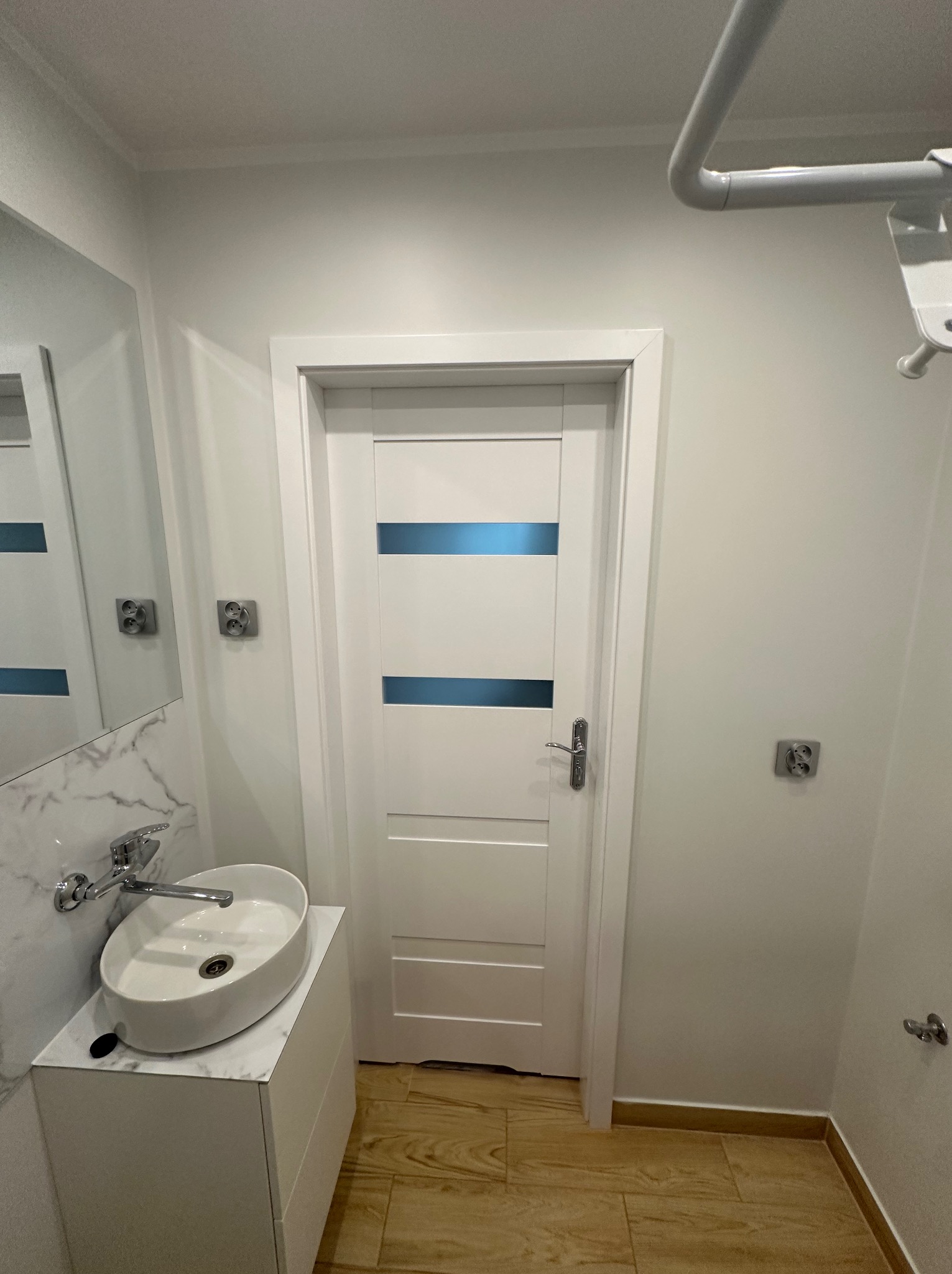 Nowoczesna łazienka z białą umywalką nablatową na szafce z blatem imitującym marmur, białe drzwi z poziomymi niebieskimi wstawkami i podłoga imitująca drewno.