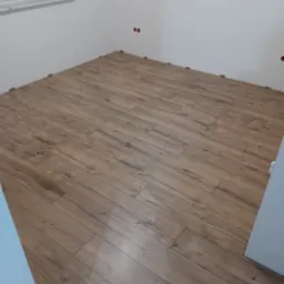 Fragment pokoju z częściowo ułożonymi panelami podłogowymi o jasnobrązowym odcieniu, widoczne plastikowe kliny dystansowe przy ścianach, surowa ściana z gniazdkami elektrycznymi i okno z folią...