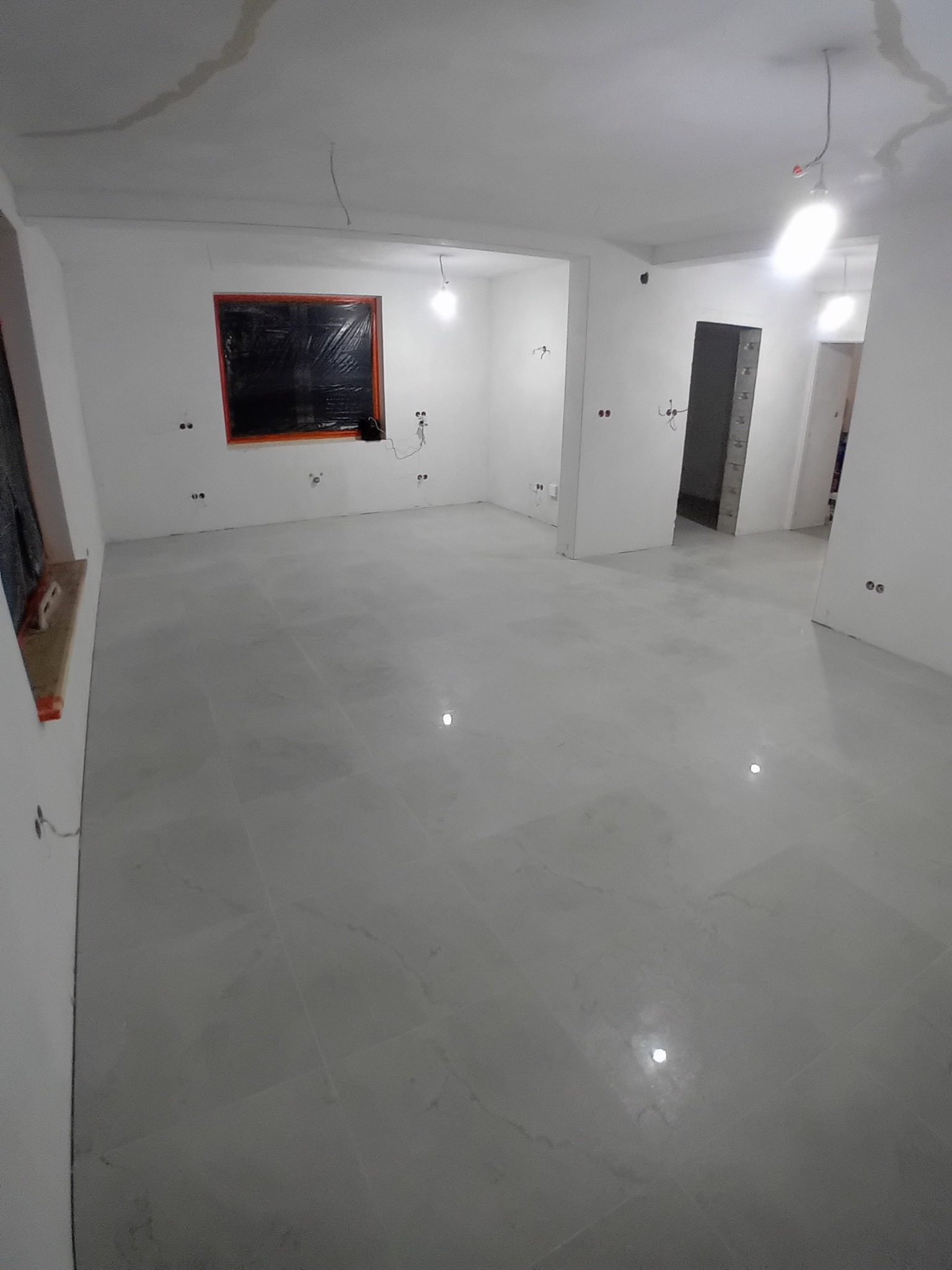 Wnętrze po remoncie z jasnoszarą, błyszczącą podłogą, zabezpieczonym oknem folią i przygotowanymi punktami elektrycznymi na białych ścianach.