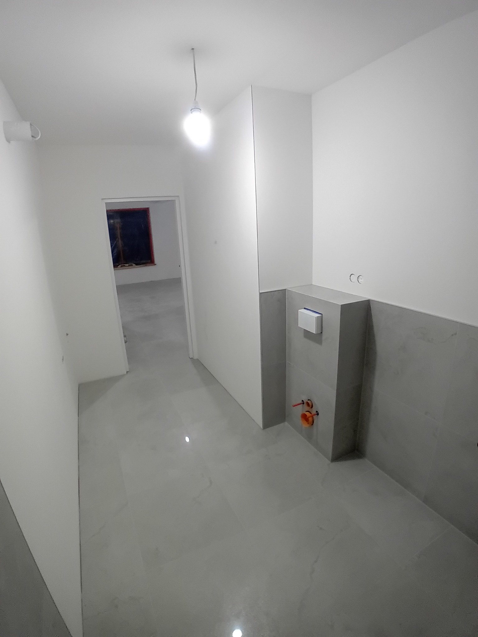 Wnętrze łazienki w trakcie remontu, widoczne szare płytki ścienne i podłogowe, zabudowa podtynkowego WC z przyciskiem, instalacja wodna z pomarańczowymi rurkami, surowa żarówka na kablu i otwarte...