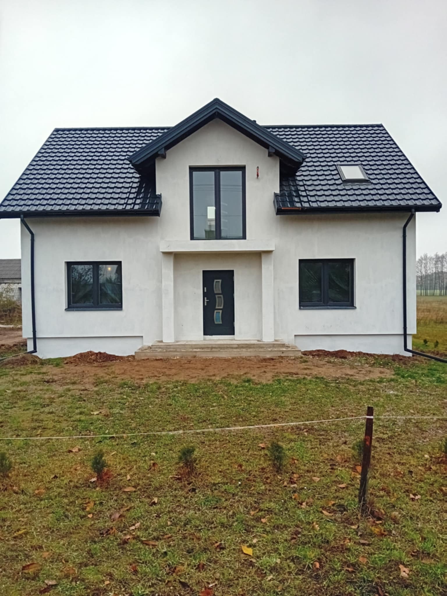 Nowoczesny dom jednorodzinny z białą elewacją, czarnym dachem z dachówki i ciemnymi oknami, widziany z przodu na tle pochmurnego nieba i trawnika.