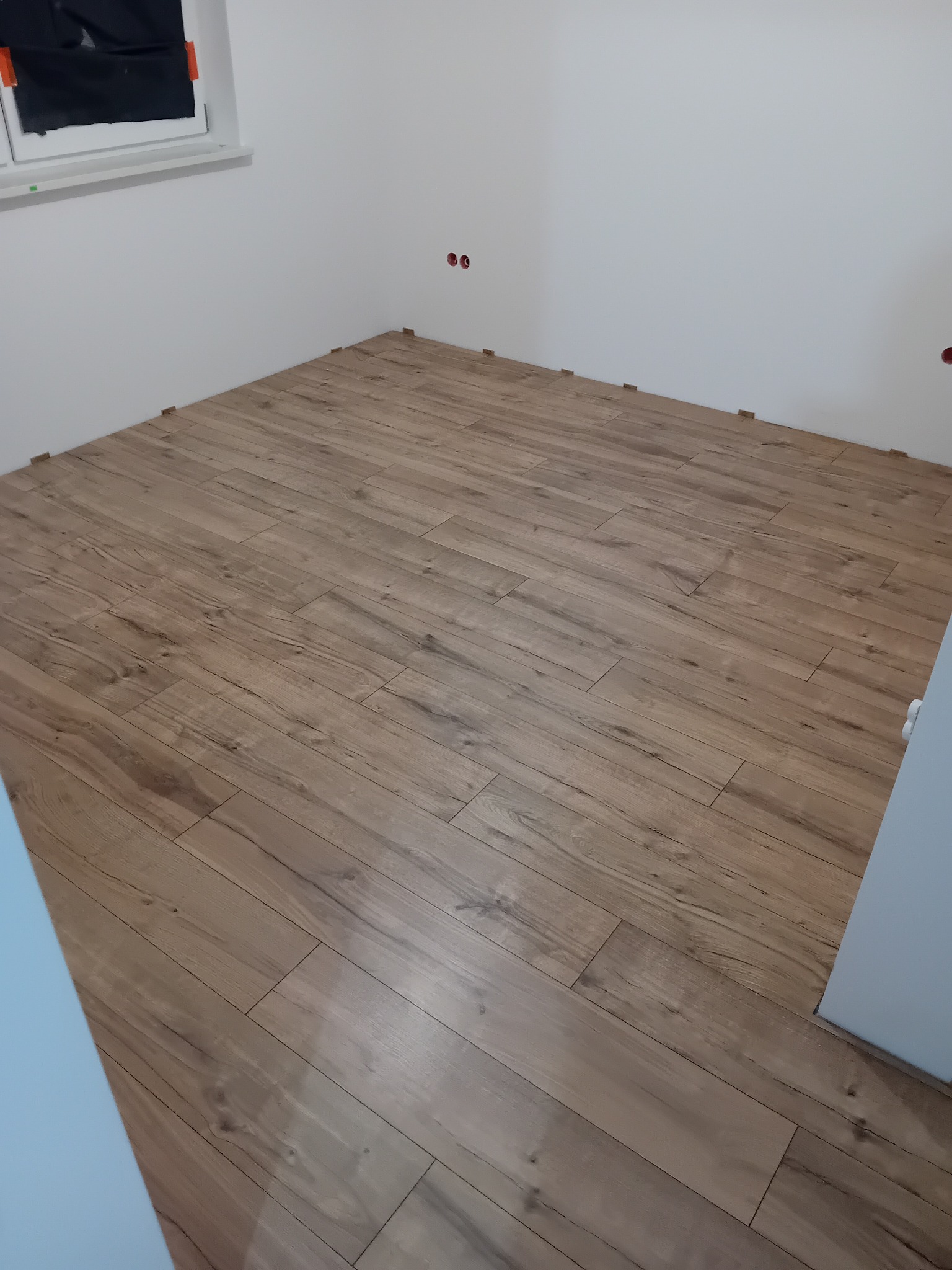 Fragment pokoju z częściowo ułożonymi panelami podłogowymi o jasnobrązowym odcieniu, widoczne plastikowe kliny dystansowe przy ścianach, surowa ściana z gniazdkami elektrycznymi i okno z folią...