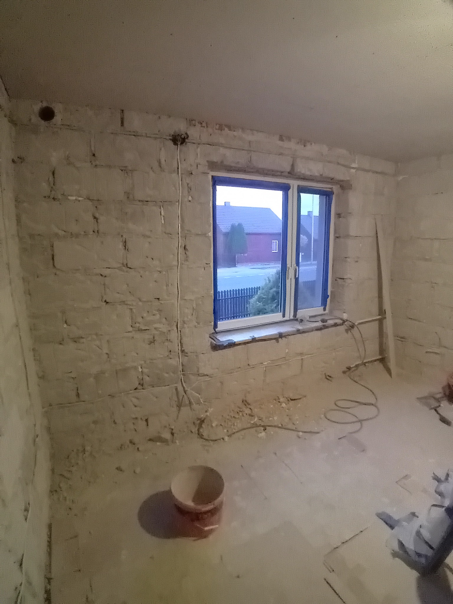 Surowe wnętrze pokoju w trakcie remontu: ściany z cegły, okno z widokiem na dom, narzędzia i wiadro na podłodze. Przygotowanie do wykończenia ścian.