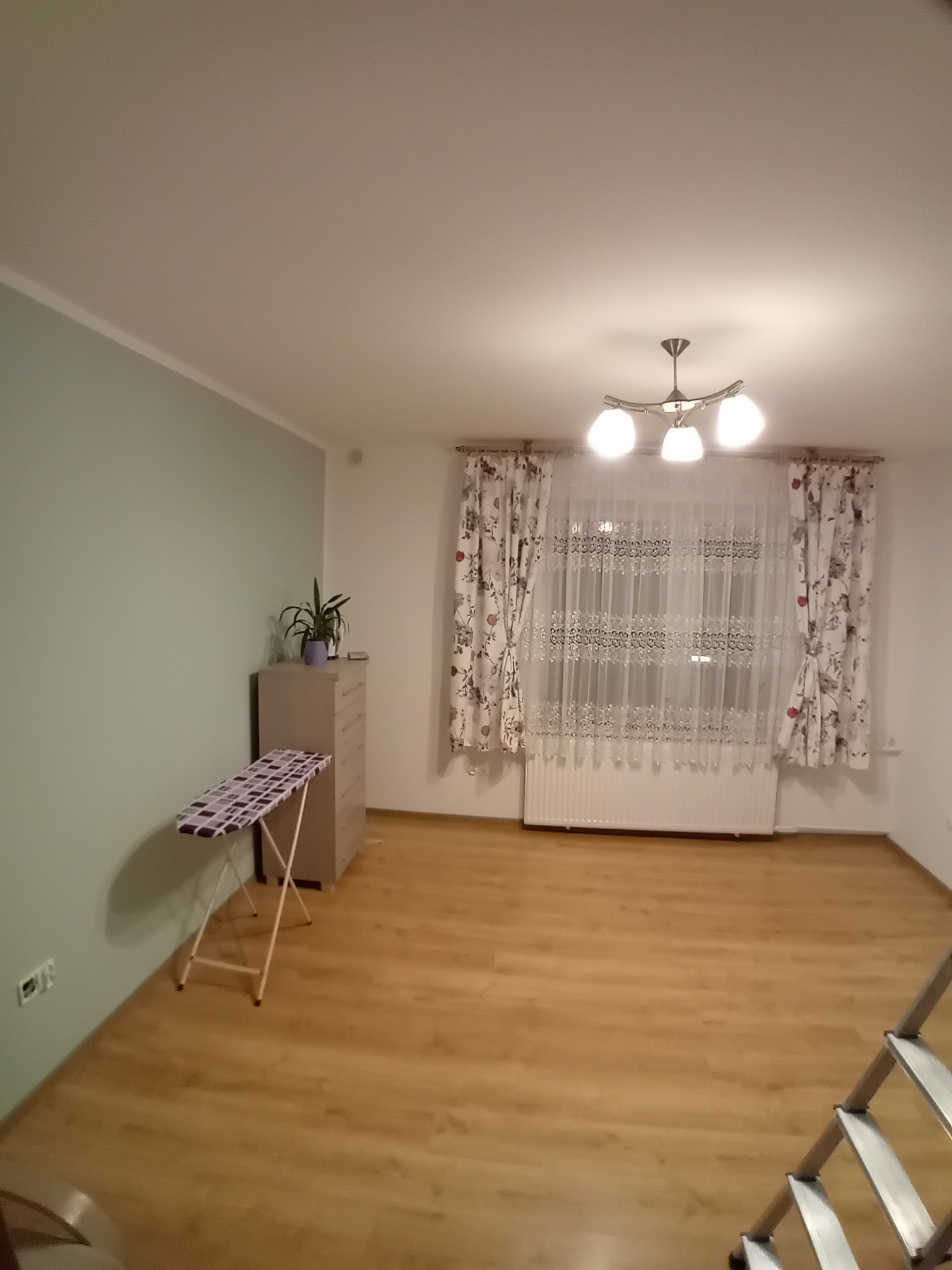 Jasne wnętrze pokoju po odświeżeniu: panele podłogowe, ściana w odcieniach zieleni, okno z firankami i zasłonami w kwiaty, komoda z rośliną i deska do prasowania.