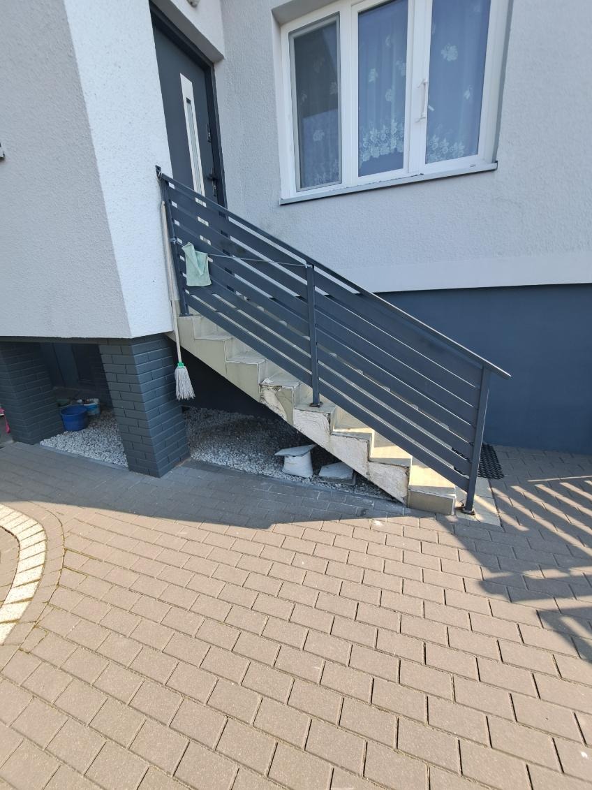 Schody wejściowe z szarą metalową barierką, prowadzące do drzwi budynku. Widoczne ślady prac remontowych, mop oparty o barierkę, kostka brukowa na pierwszym planie.