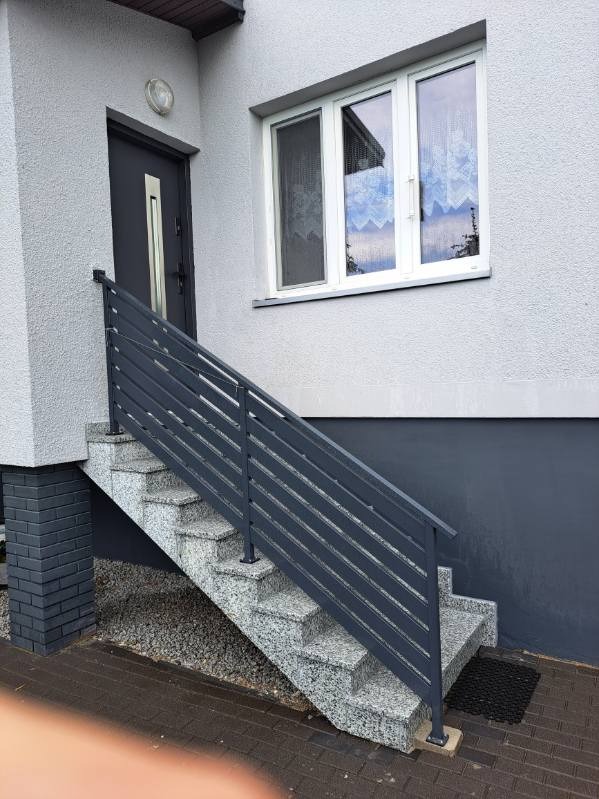 Schody wejściowe z szarego granitu z metalową, poziomą balustradą. Elewacja budynku w odcieniach szarości, okno z białymi ramami. Widok z dołu, lekko z boku.