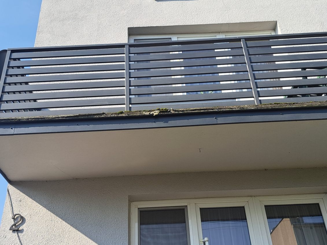 Szary balkon z poziomymi szczebelkami, widok z dołu, z numerem '2' na ścianie budynku. Widoczne okna i fragment nieba.