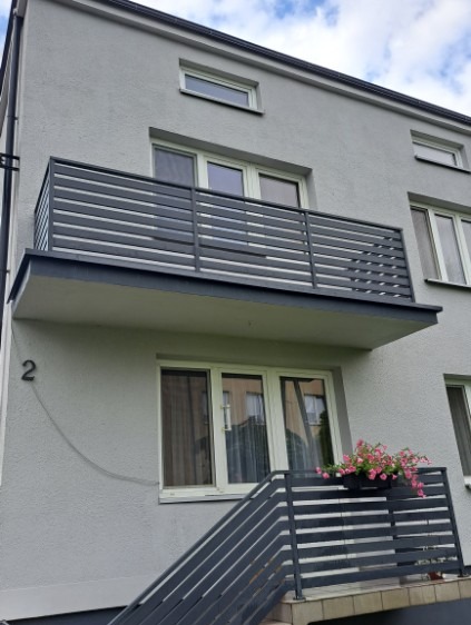 Odnowiony balkon z szarą, poziomą balustradą w bloku mieszkalnym. Kwiaty w doniczce na balustradzie schodów. Numer '2' na elewacji. Jasna elewacja budynku.
