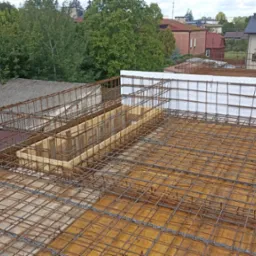 Zbrojenie stropu z drewnianym szalunkiem i białym styropianem, widoczne z góry na tle zieleni i zabudowy miejskiej. Etap prac budowlanych z użyciem metalowych prętów.