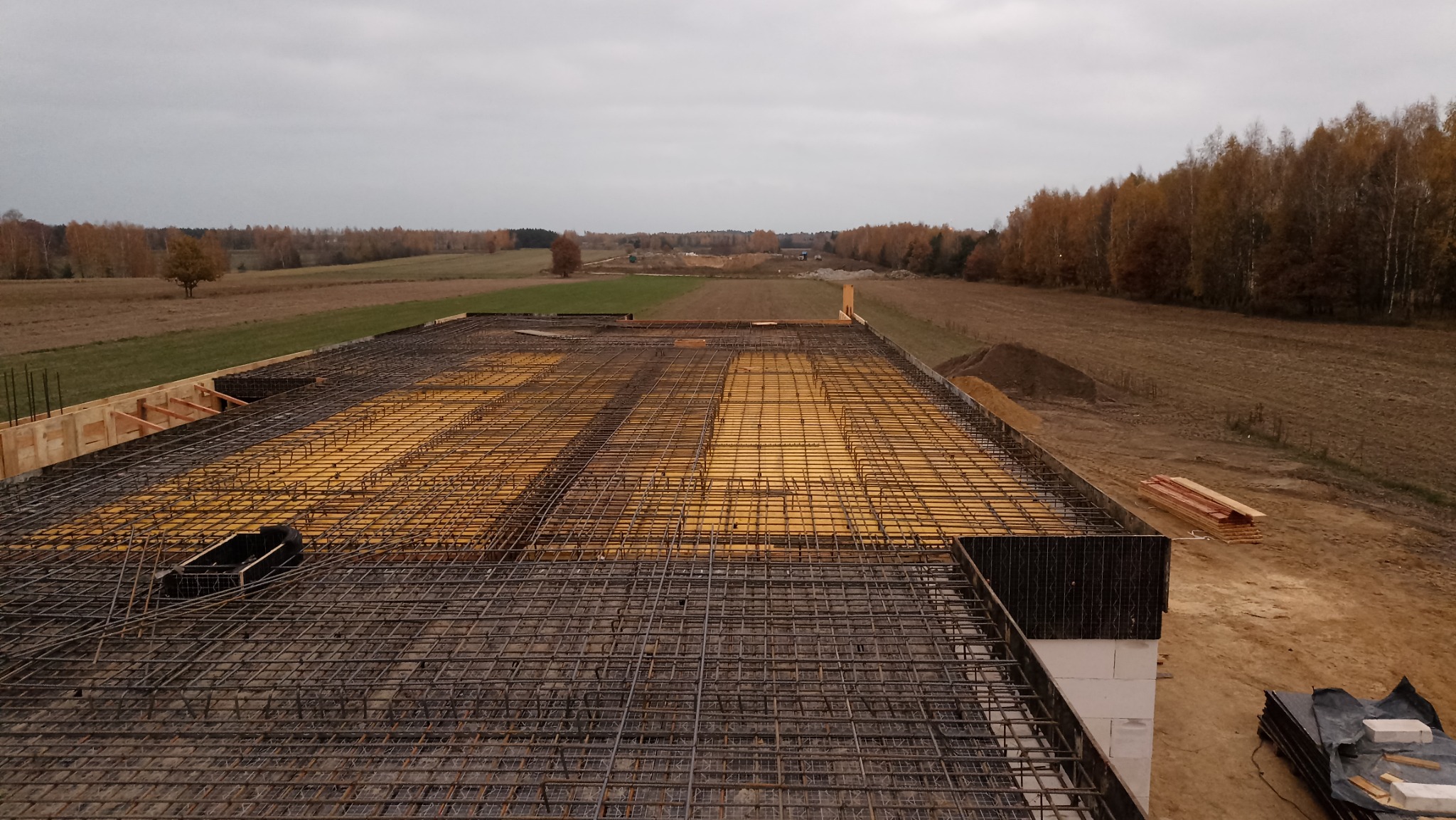 Zbrojenie stropu domu murowanego z widocznym deskowaniem i siatką prętów stalowych na tle jesiennego krajobrazu.