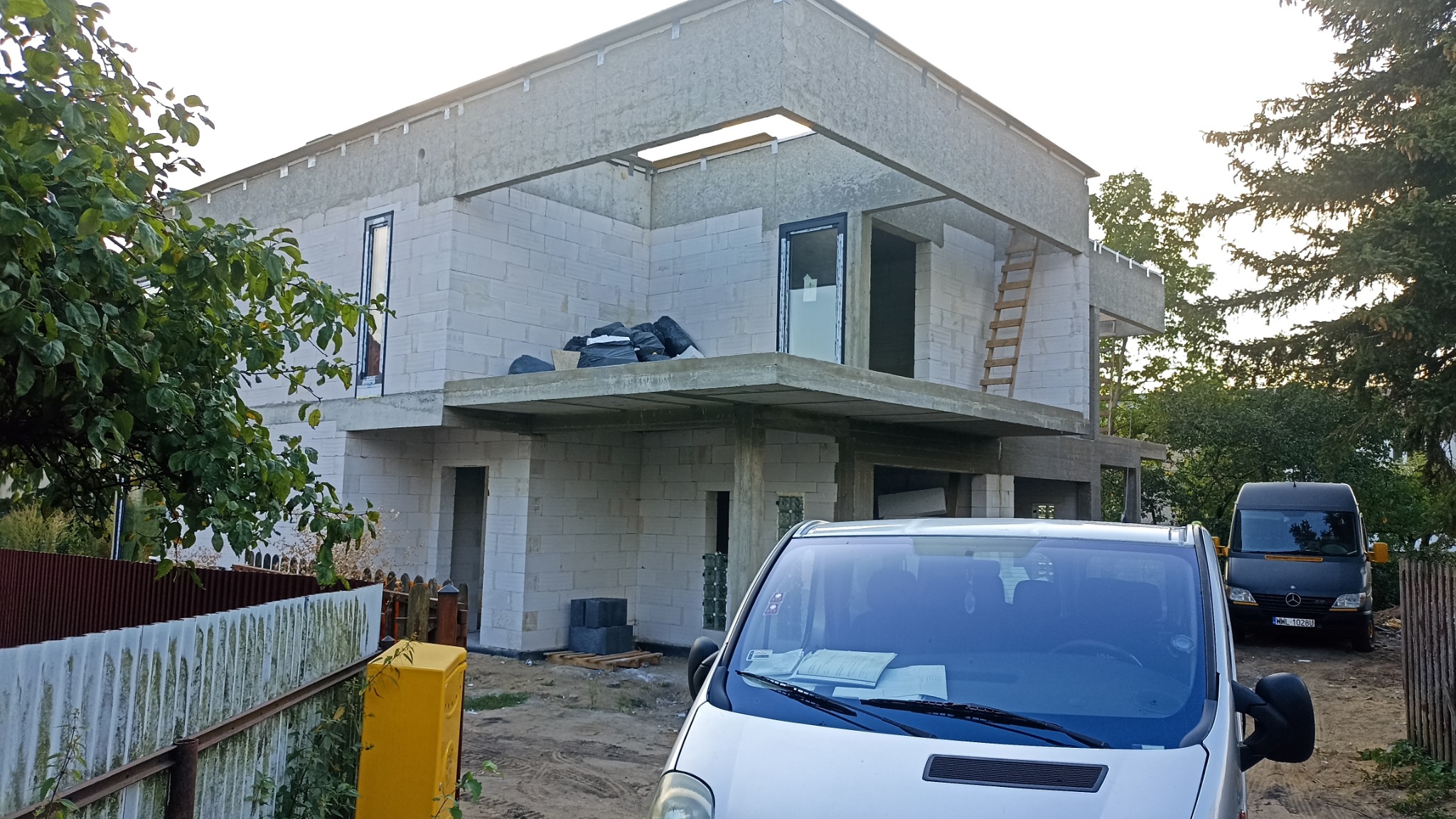 Surowy, dwukondygnacyjny dom z betonu komórkowego w trakcie budowy, z widocznymi otworami okiennymi i czarnymi workami na balkonie. Dwa samochody dostawcze na posesji.