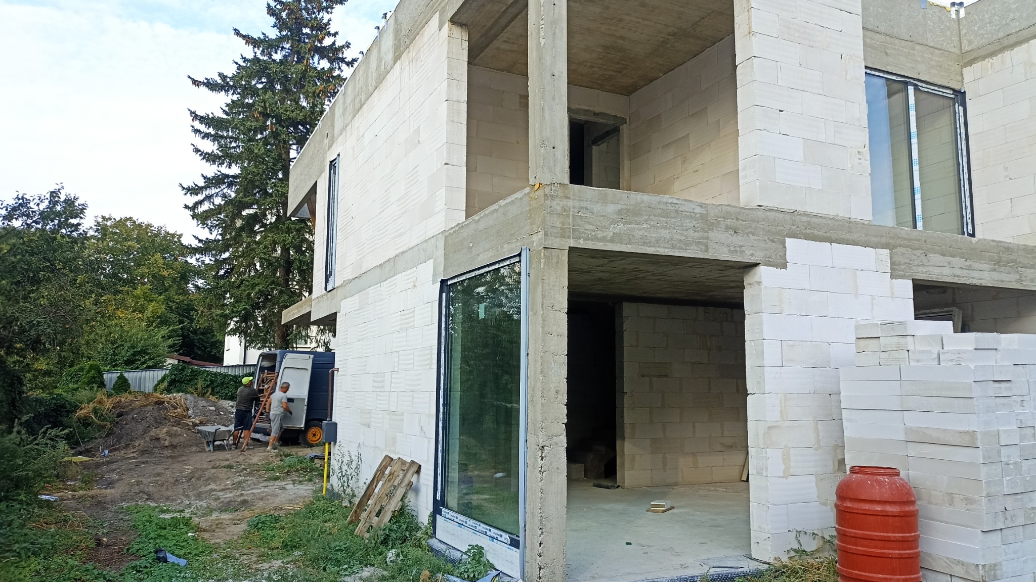 Surowy stan dwukondygnacyjnego domu z betonu komórkowego w trakcie budowy, z widocznymi oknami i stertą bloczków na zewnątrz, z pracownikami w tle.