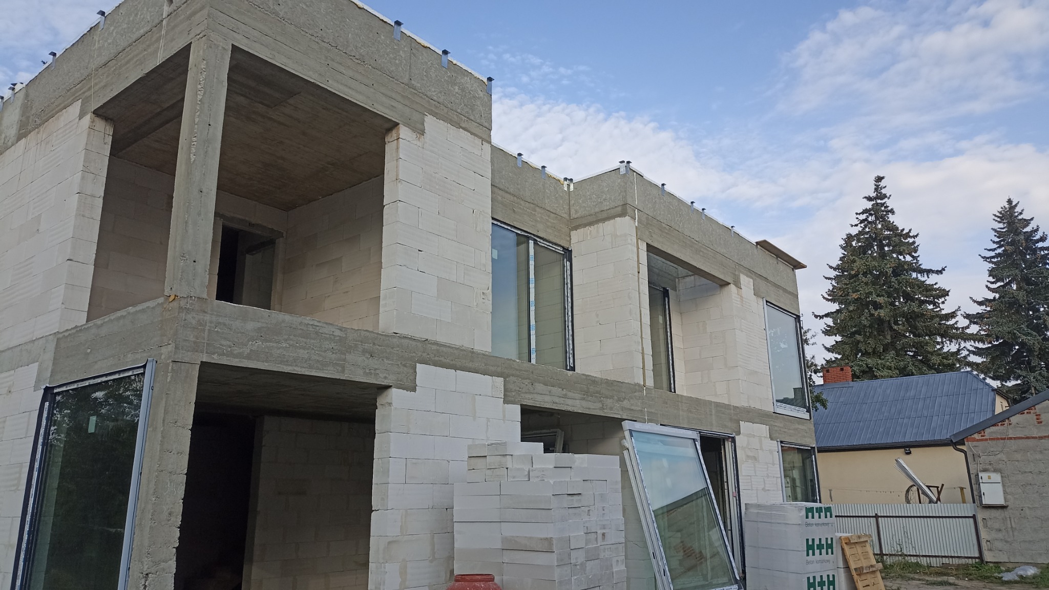 Surowy stan dwukondygnacyjnego domu z betonu komórkowego, z wstawionymi oknami. Na pierwszym planie paleta z bloczkami i okno. W tle budynek sąsiada.