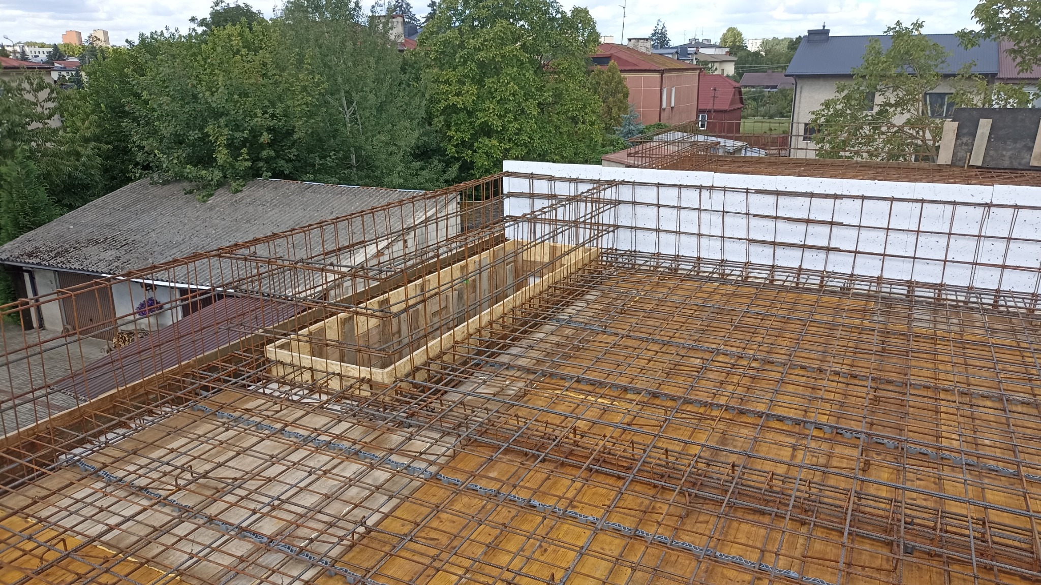 Zbrojenie stropu z drewnianym szalunkiem i białym styropianem, widoczne z góry na tle zieleni i zabudowy miejskiej. Etap prac budowlanych z użyciem metalowych prętów.