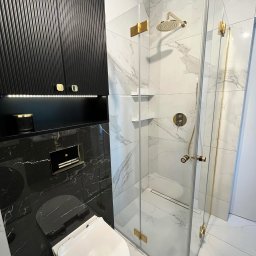 Przemysław Rychlik PHU Rychmal - Nowoczesna łazienka z kabiną prysznicową z elementami w kolorze złotym, wiszącą toaletą na czarnej marmurowej ścianie i czarną szafką z ryflowanymi drzwiczkami.