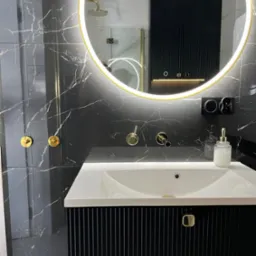 Nowoczesna łazienka z czarnymi marmurowymi płytkami, okrągłym lustrem z podświetleniem LED, białą umywalką nablatową i czarną szafką z ryflowanym frontem oraz złotymi akcentami w postaci armatury...