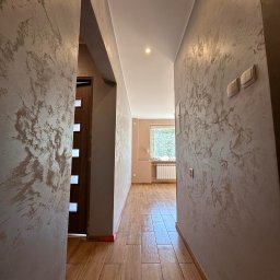 Przemysław Rychlik PHU Rychmal - Korytarz po odświeżeniu, z dekoracyjną fakturą na ścianach w odcieniach beżu i brązu, drewnopodobna podłoga z płytek ceramicznych, widok na jasny pokój z grzejnikiem pod oknem.