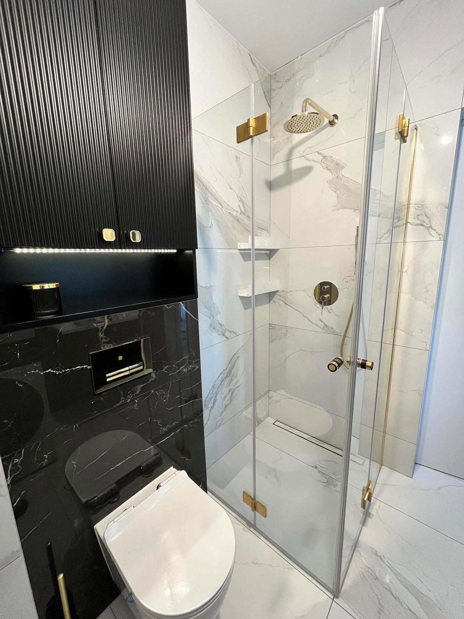 Nowoczesna łazienka z kabiną prysznicową z elementami w kolorze złotym, wiszącą toaletą na czarnej marmurowej ścianie i czarną szafką z ryflowanymi drzwiczkami.