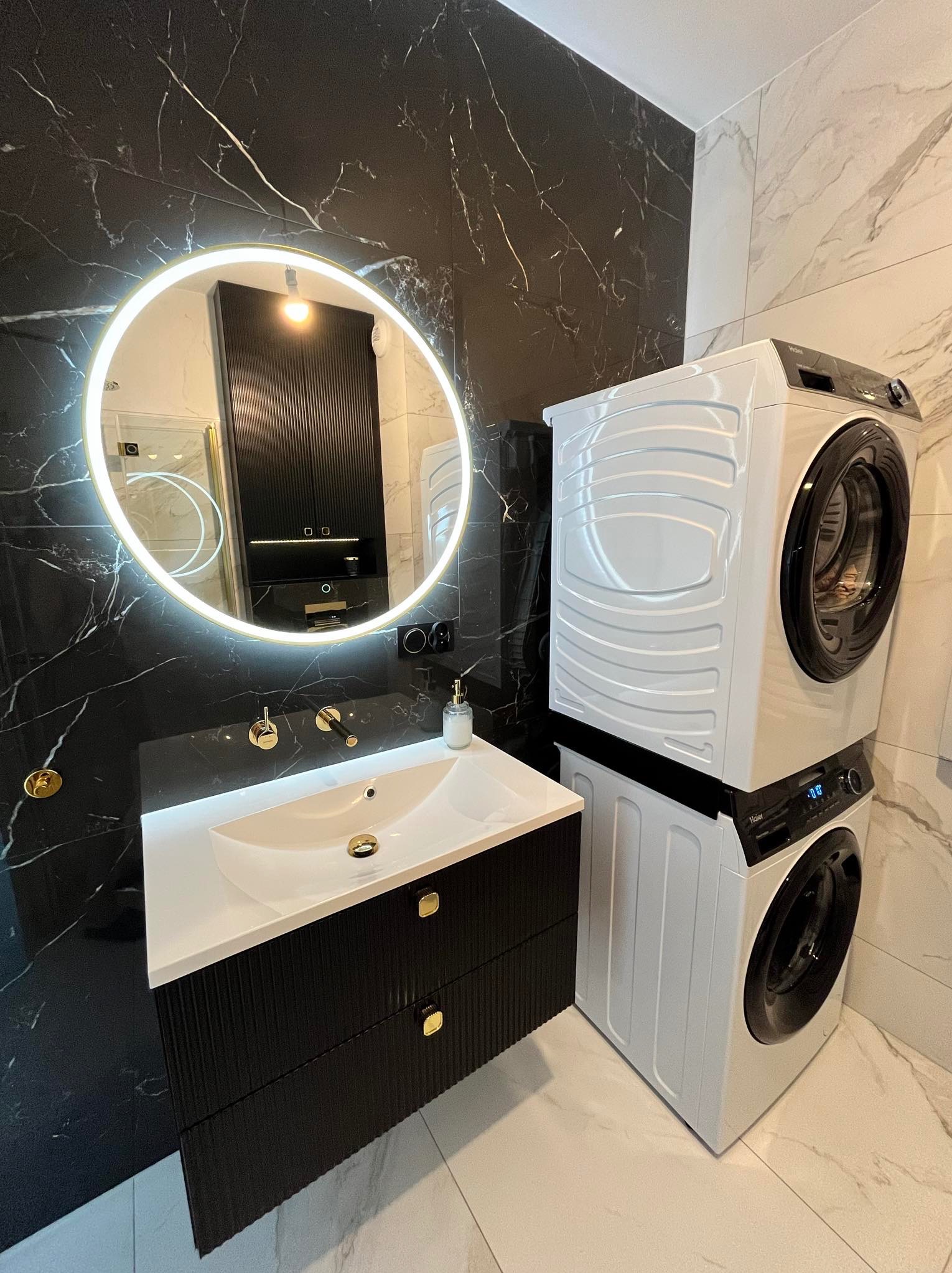Nowoczesna łazienka w Pleszewie z czarnymi marmurowymi płytkami, podświetlanym okrągłym lustrem nad białą umywalką z czarną szafką i złotymi detalami oraz pralką i suszarką ustawioną jedna na drugiej.