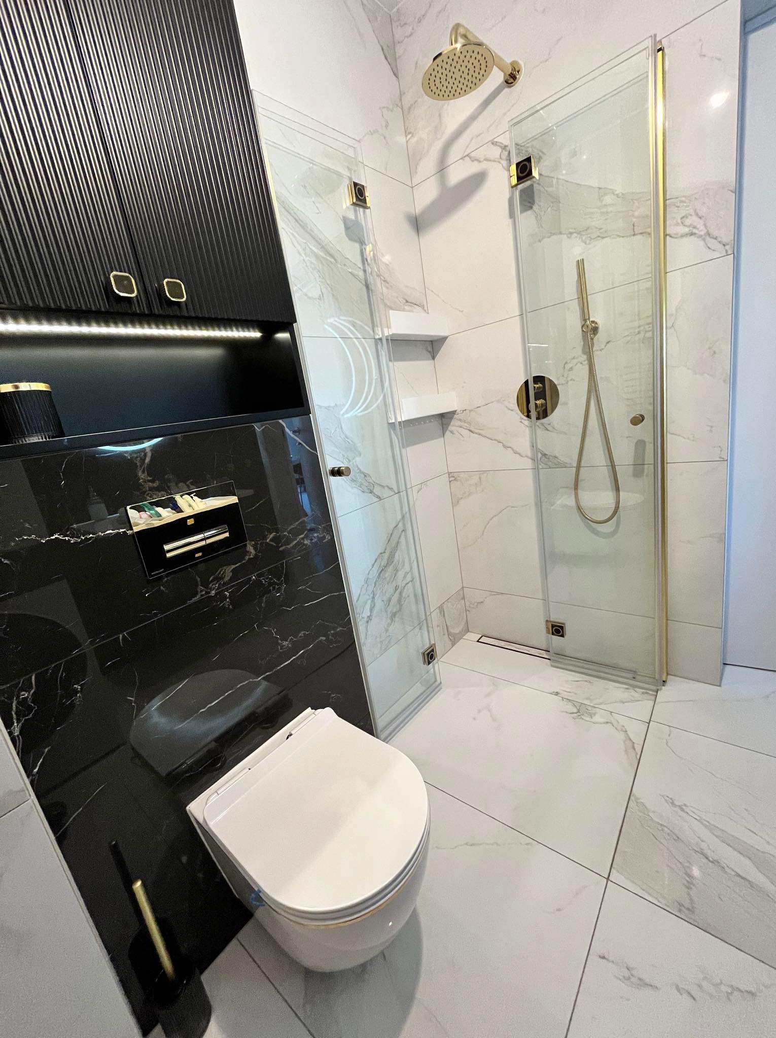 Nowoczesna łazienka z czarnymi szafkami z ryflowanymi frontami, białą ceramiką i kabiną prysznicową ze złotymi okuciami na tle płytek imitujących marmur.