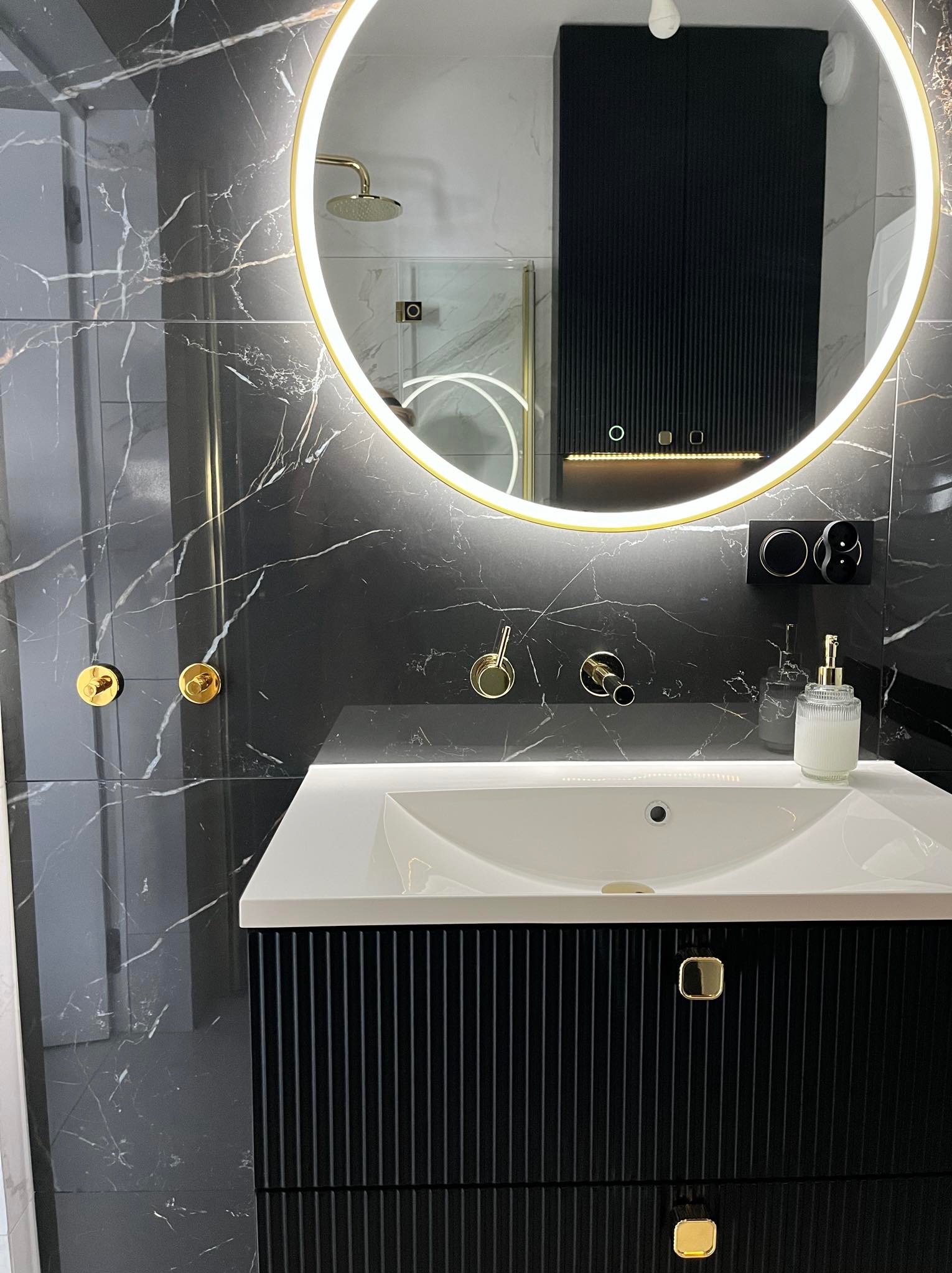 Nowoczesna łazienka z czarnymi marmurowymi płytkami, okrągłym lustrem z podświetleniem LED, białą umywalką nablatową i czarną szafką z ryflowanym frontem oraz złotymi akcentami w postaci armatury...