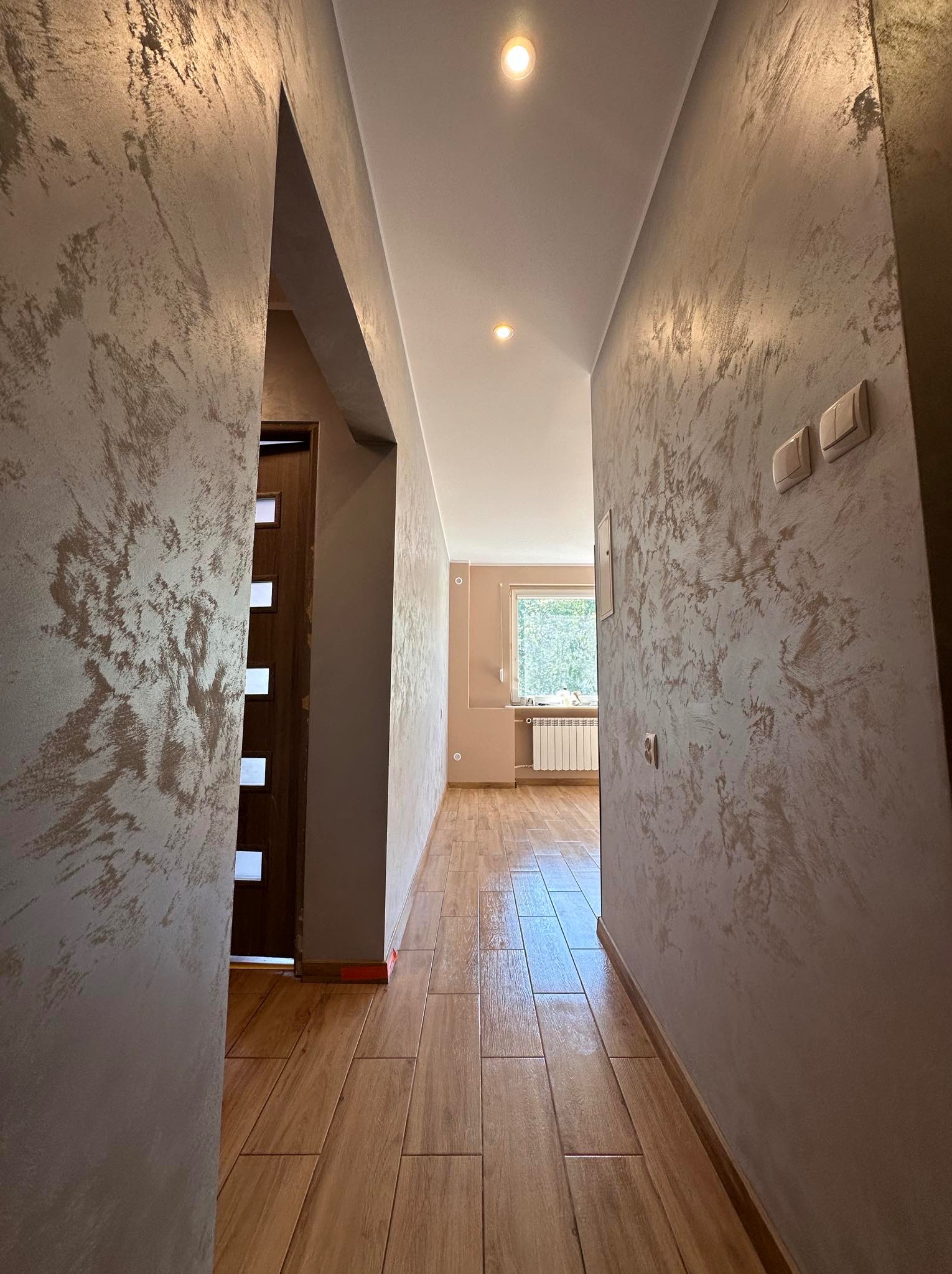 Korytarz po odświeżeniu, z dekoracyjną fakturą na ścianach w odcieniach beżu i brązu, drewnopodobna podłoga z płytek ceramicznych, widok na jasny pokój z grzejnikiem pod oknem.