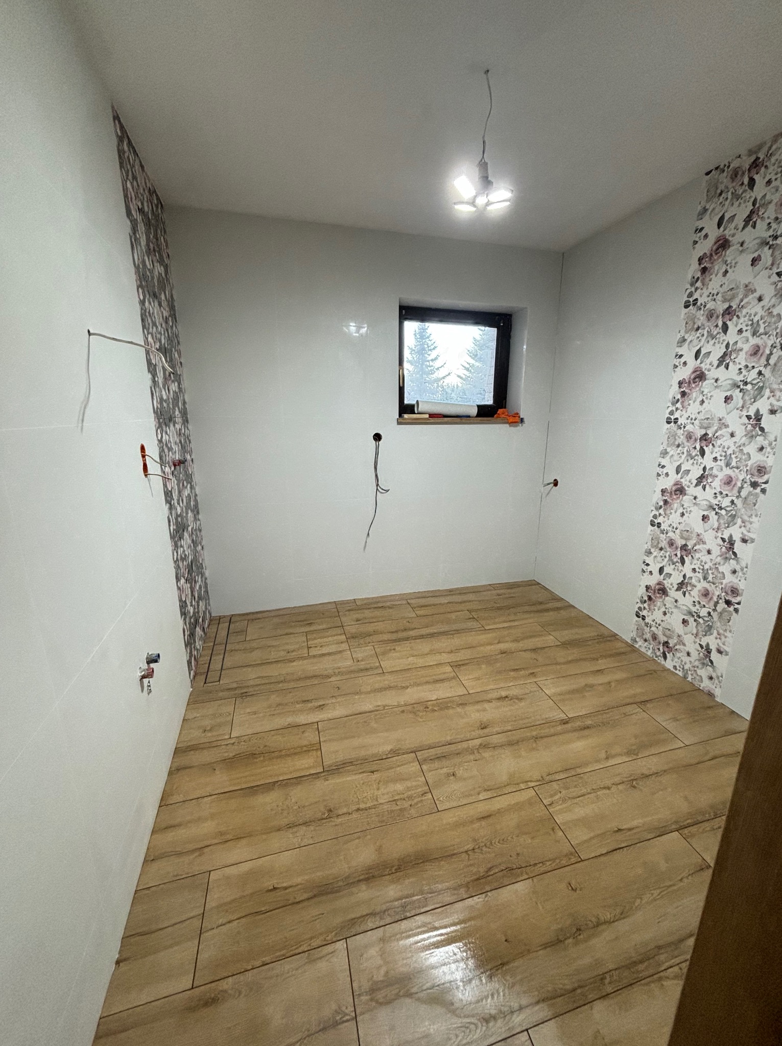 Wnętrze łazienki w trakcie prac: ułożona podłoga z płytek imitujących drewno, częściowo położona tapeta w kwiaty na ścianach, widoczne wystające kable i rury, okno z widokiem na zaśnieżone drzewa.