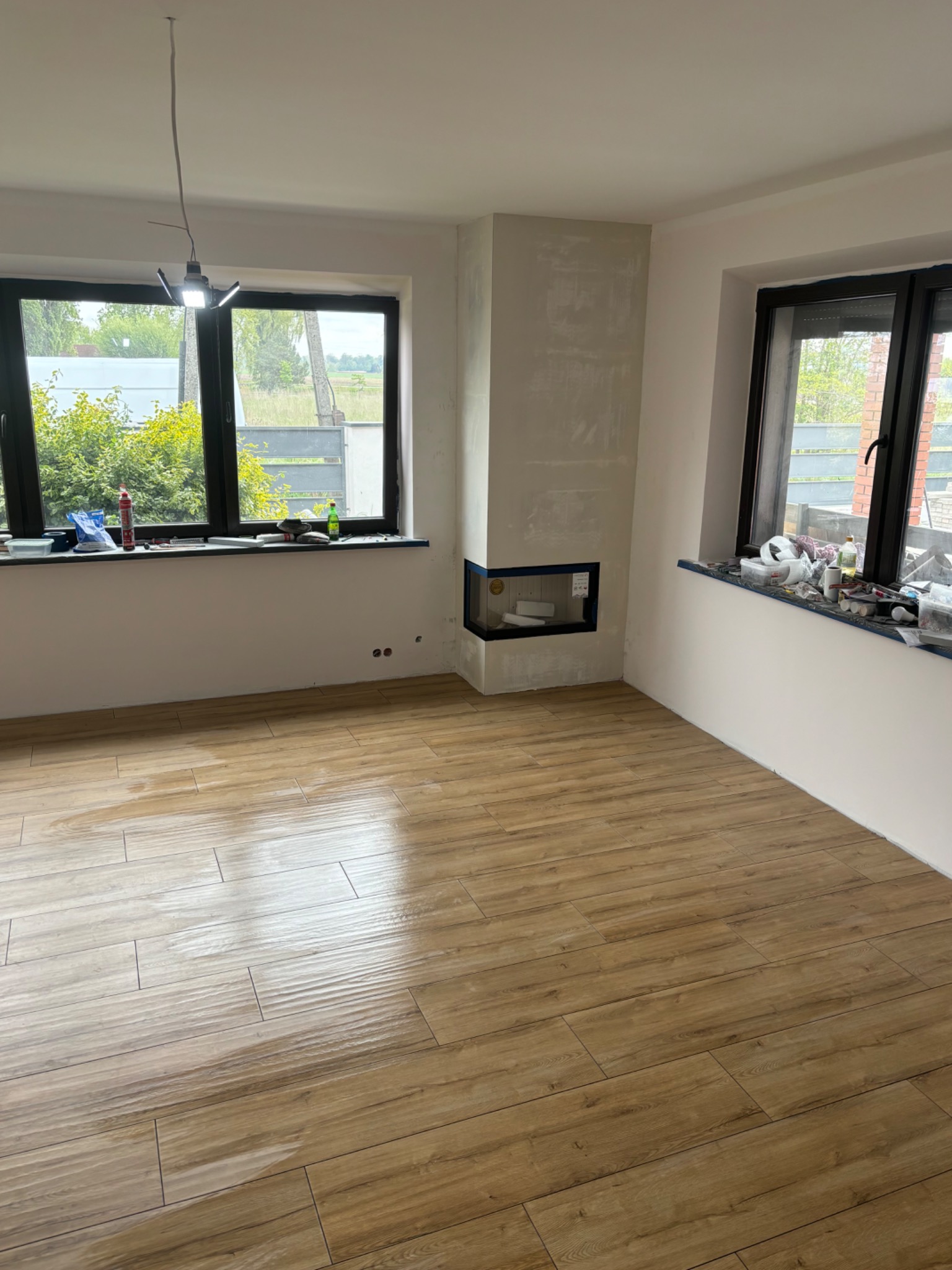 Wnętrze pokoju z nową, drewnopodobną podłogą, przygotowane do dalszych prac wykończeniowych, widoczny komin z surowym wykończeniem i narzędzia na parapecie.