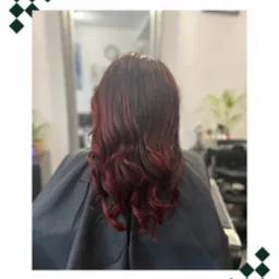 Widok z tyłu na ciemne, pofalowane włosy z czerwonymi refleksami, klientka w czarnej pelerynie fryzjerskiej, w tle lustro i wyposażenie salonu.