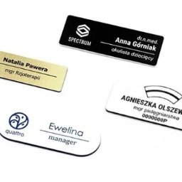 Strefaprezentacji.pl - Zestaw czterech identyfikatorów imiennych w różnych stylach: czarny z logo Spectrum, złoty szczotkowany dla mgr fizjoterapii, biały z logo Quattro dla managera, oraz biały z ozdobnym paskiem...