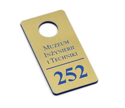 Złota zawieszka na drzwi z niebieskim napisem 'Muzeum Inżynierii i Techniki' oraz numerem 252, zawieszka z otworem na górze, leżąca ukośnie na białym tle.