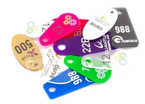 Kolorowe, plastikowe identyfikatory z nadrukowanymi numerami i logo firm na białym tle, widoczne różne kształty i kolory: niebieski, zielony, fioletowy, różowy, biały, czarny i beżowy.