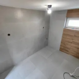 Ukończona łazienka z szarymi płytkami wielkoformatowymi na ścianach i podłodze, drewnopodobne płytki wokół okna, surowa żarówka. Widok na narożnik.