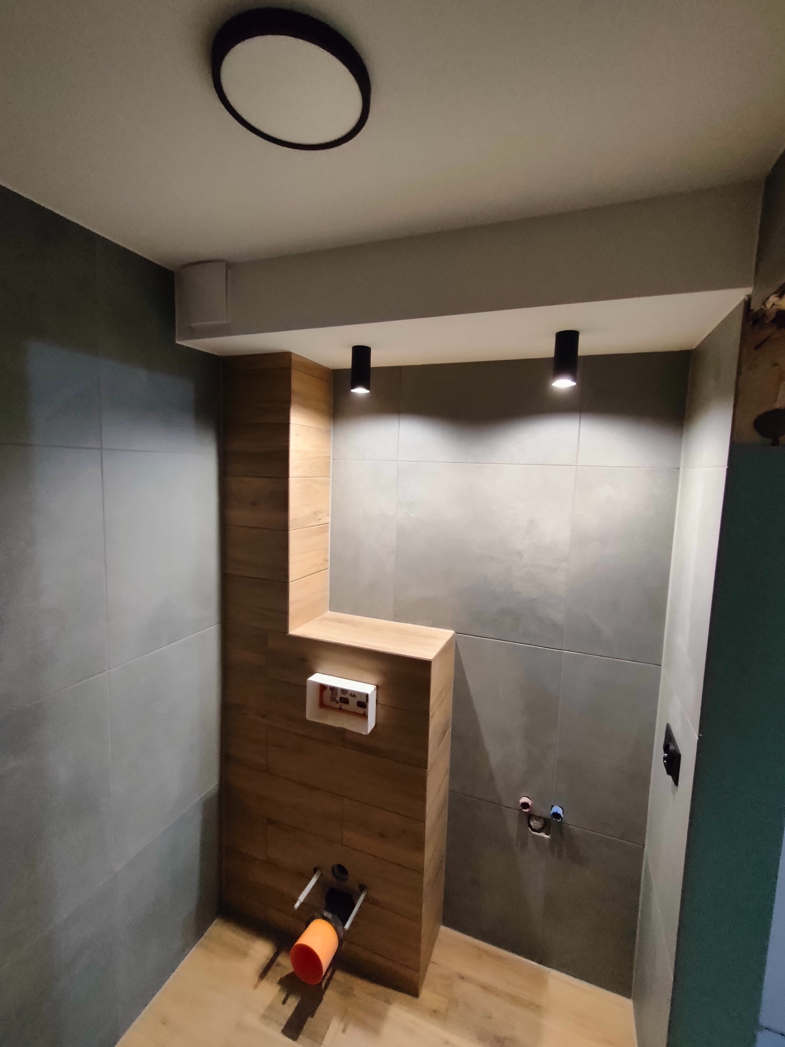 Stelaż podtynkowy WC obudowany płytkami imitującymi drewno, szare płytki na ścianach, widoczne przyłącza wodne, sufit z oświetleniem punktowym, faza montażu.