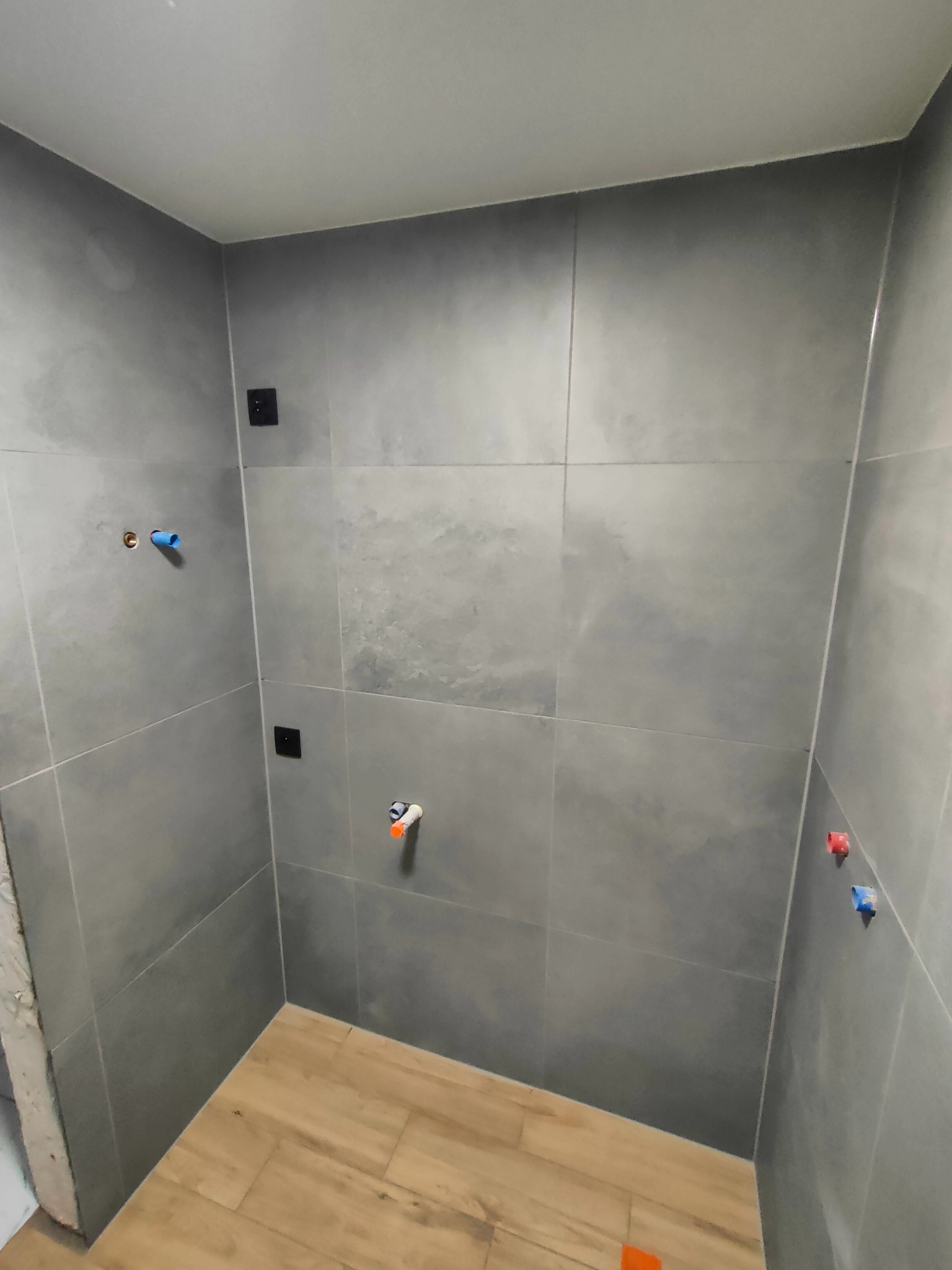 Wnętrze łazienki w trakcie prac wykończeniowych: szare, matowe płytki na ścianach, imitacja drewna na podłodze, widoczne przyłącza wodne i elektryczne, fragment surowej ściany przy wejściu.