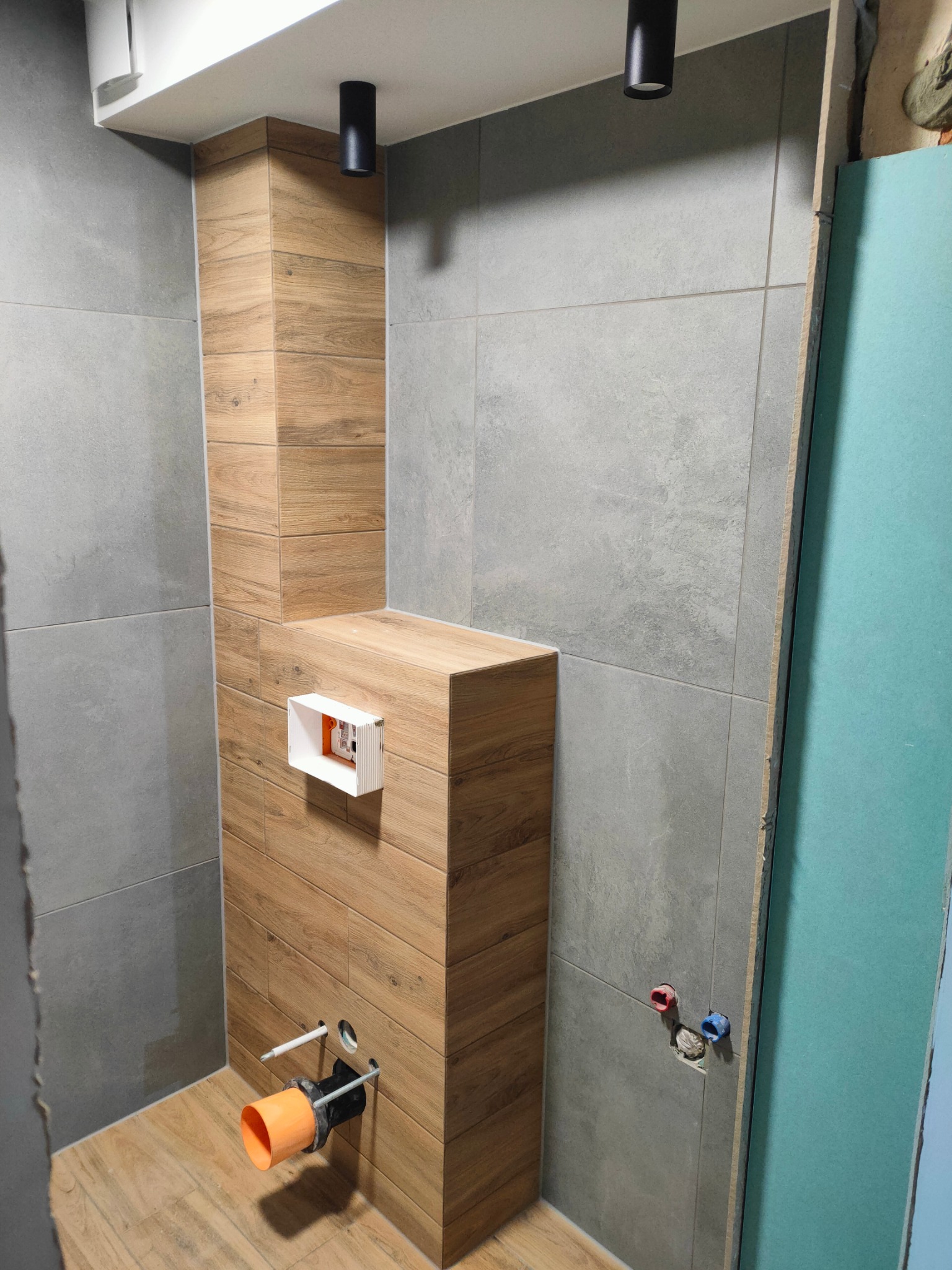Wnętrze łazienki w trakcie prac wykończeniowych: obudowa stelaża WC z płytek imitujących drewno, szare płytki na ścianach, widoczne instalacje wodno-kanalizacyjne i płyta gipsowo-kartonowa na ścianie.