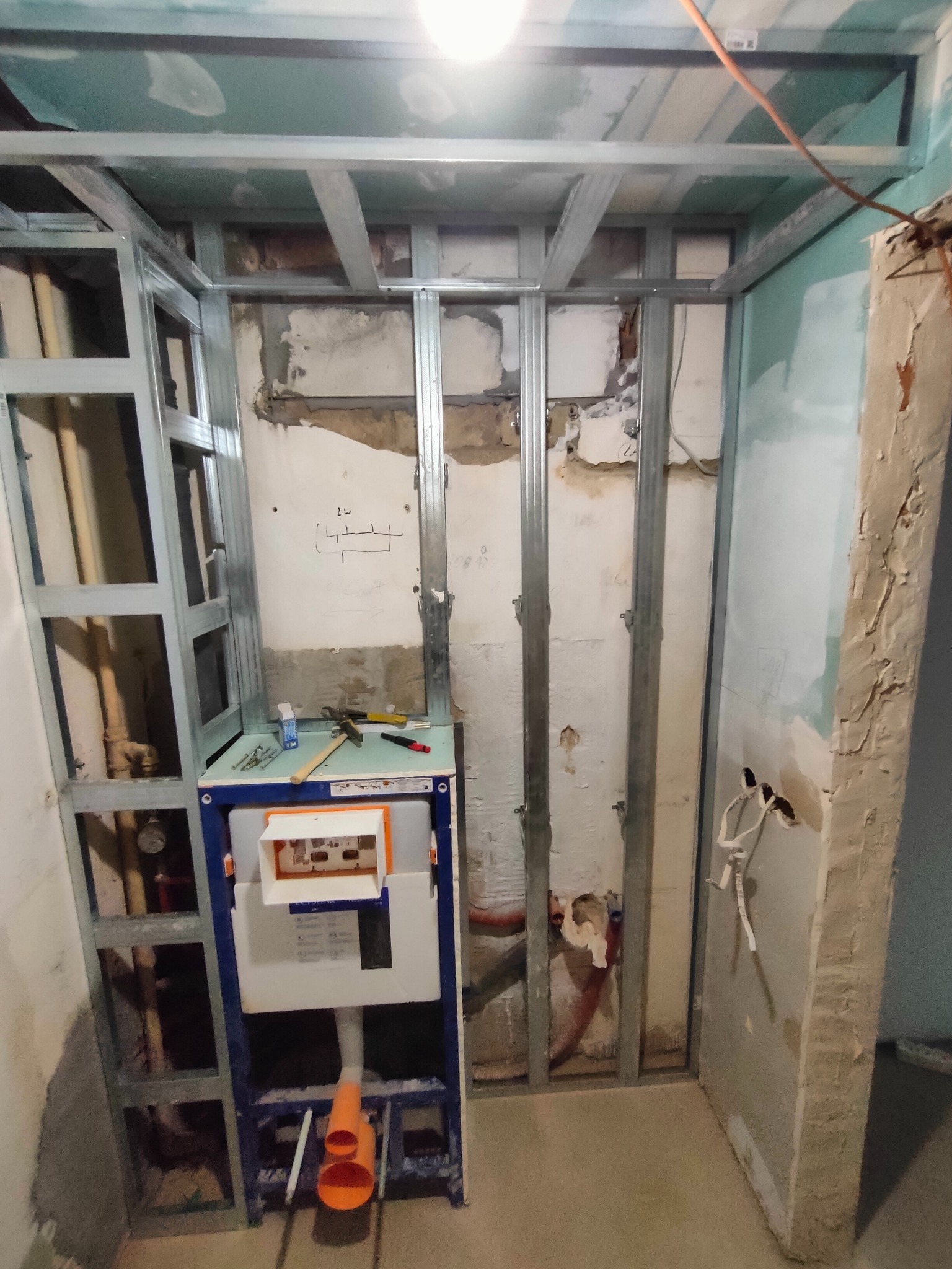 Stelaż podtynkowy WC zamontowany w metalowej konstrukcji szkieletowej, widoczne narzędzia na górnej półce, fragmenty instalacji wodnej i kanalizacyjnej, przygotowanie do obudowy ściany.