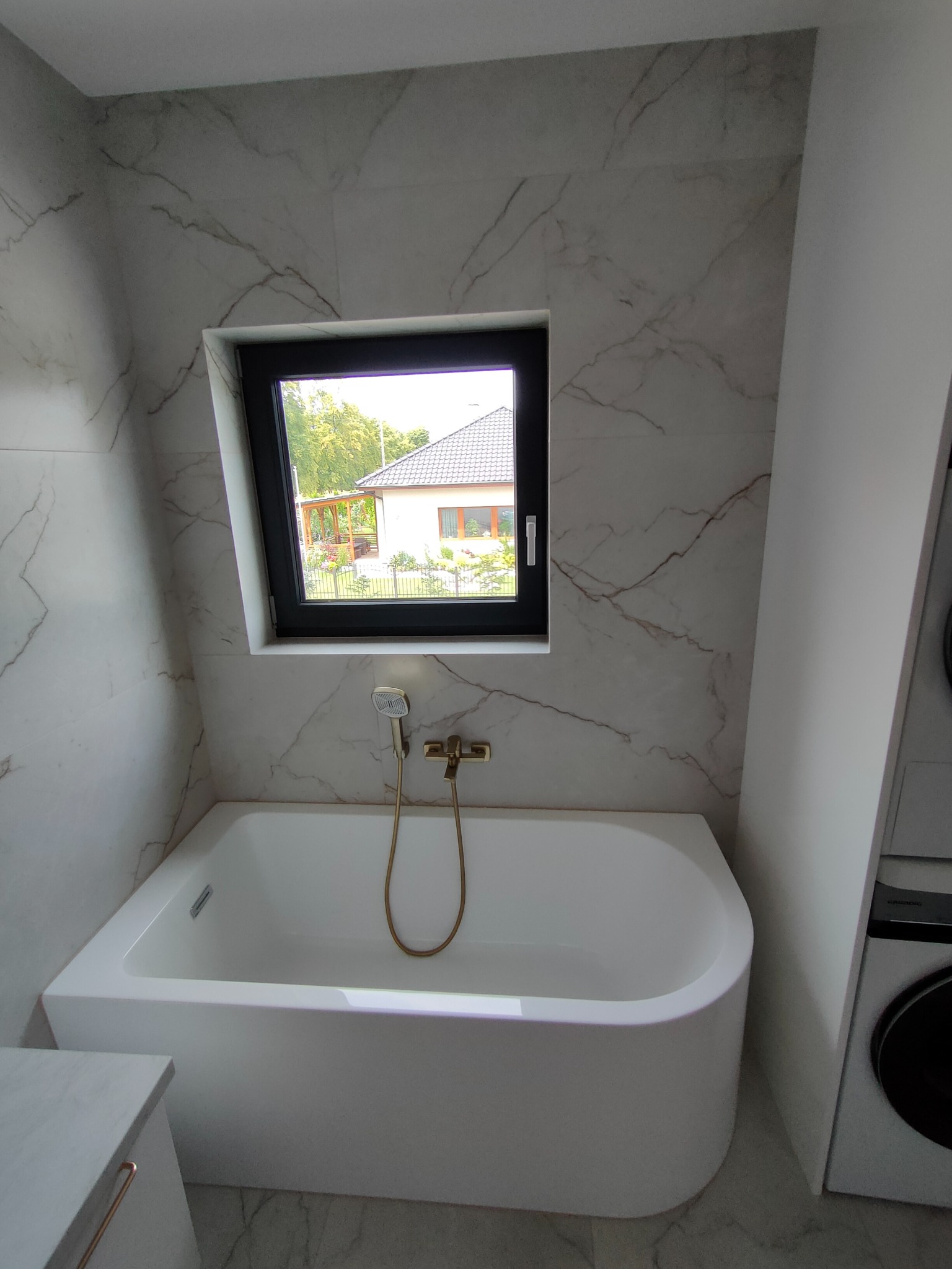 Biała wanna pod oknem w łazience wykończonej płytkami imitującymi marmur. Złota bateria i słuchawka prysznicowa. Widok z okna na dom i zieleń.